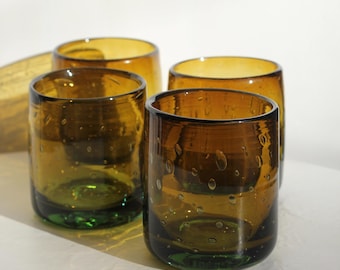 May include: Four cylindrical drinking glasses. The glasses are a transparent amber colour with a green base. The glass has small bubbles throughout. The glasses are on a white surface.