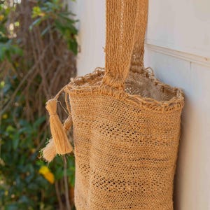 Puede incluir: Un bolso cilíndrico de fibras trenzadas en color beige, con una sola correa para el hombro y una borla decorativa en el lateral. El bolso presenta una textura de tejido abierto con un borde superior reforzado y una base circular plana.