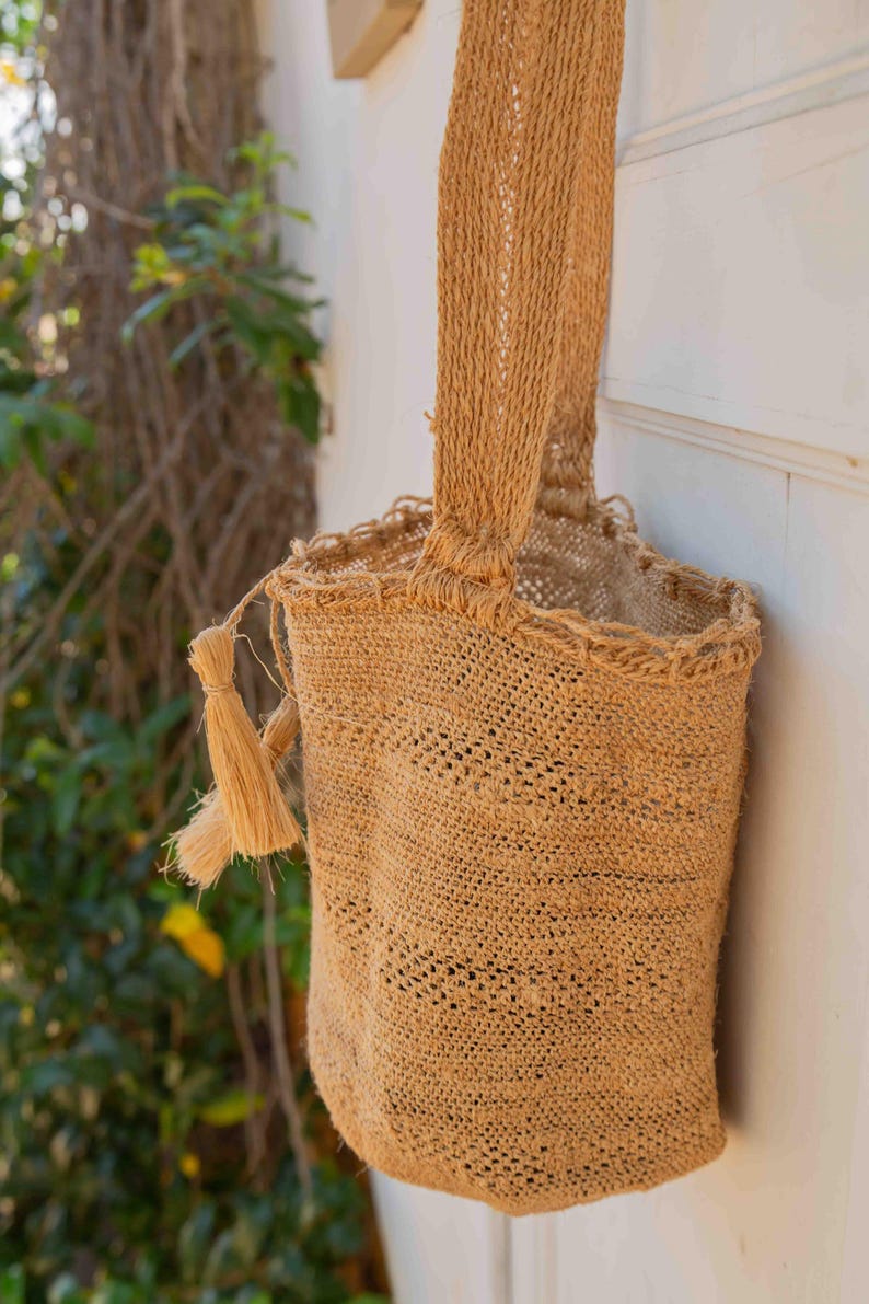 May include: A tan, cylindrical, woven fiber bag featuring a single shoulder strap and a decorative tassel attached to the side. The bag has a textured, open-weave construction with a reinforced top rim and a flat, circular base for stability.