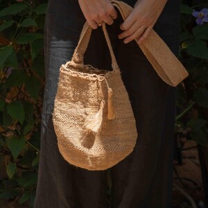 May include: A tan woven bucket bag made of natural fiber, featuring a round base, a single shoulder strap, and a drawstring closure adorned with two small matching tassels hanging from the side of the opening, showcasing a textured weave.