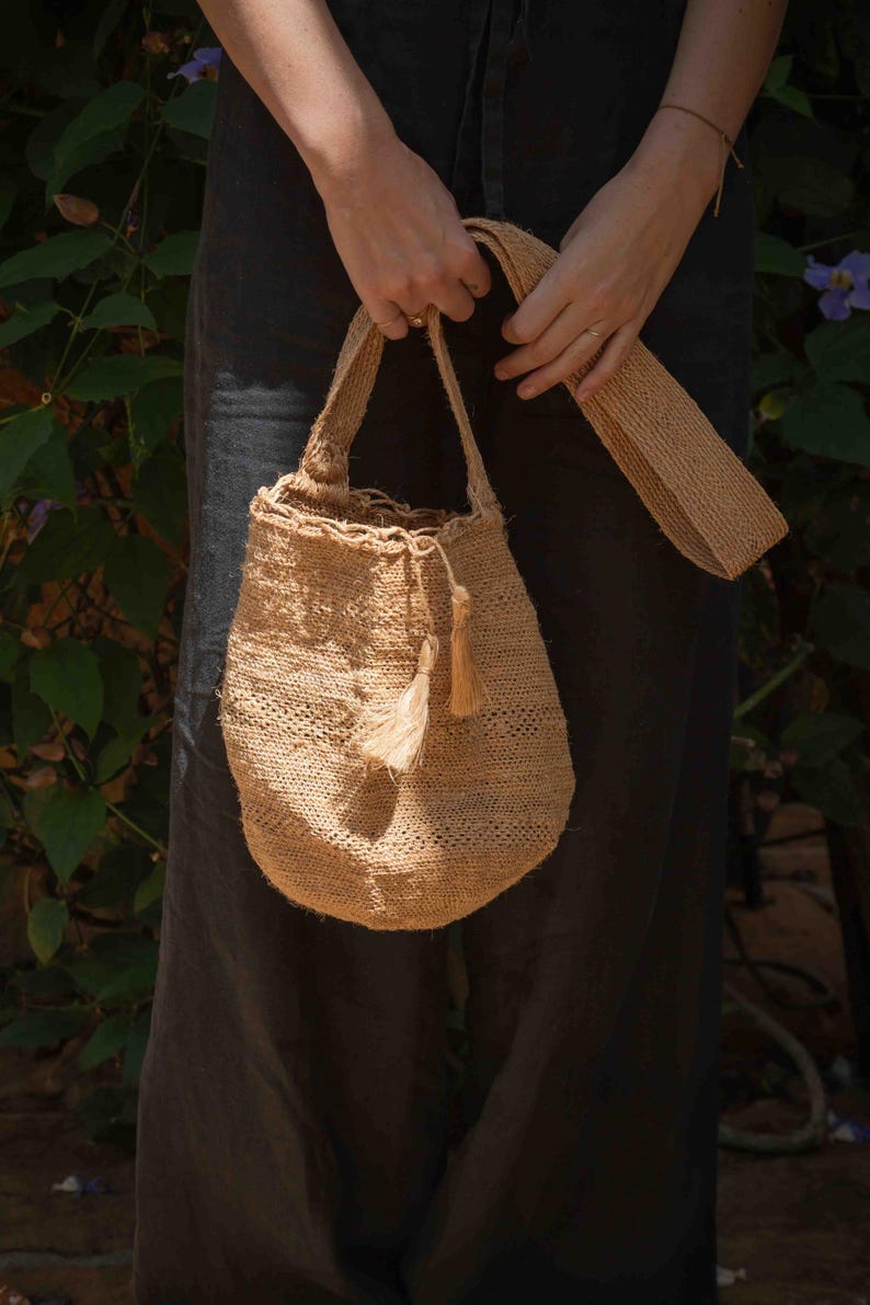 May include: A tan woven bucket bag made of natural fiber, featuring a round base, a single shoulder strap, and a drawstring closure adorned with two small matching tassels hanging from the side of the opening, showcasing a textured weave.