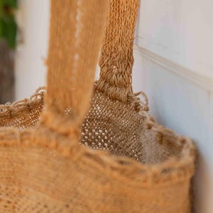 May include: A tan-colored tote bag crafted from a coarse, woven natural fiber material. The bag features a wide, sturdy handle attached to the top rim, with a visible open-weave texture throughout the body and a reinforced stitched seam at the handle.