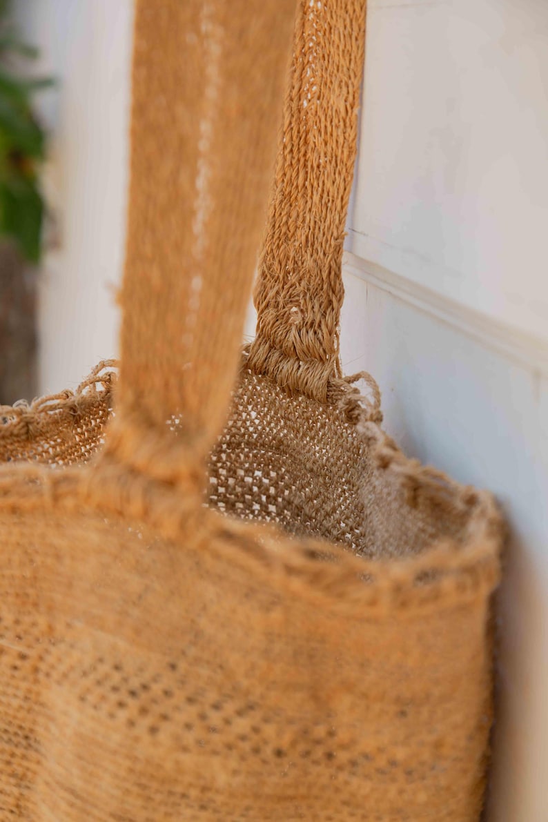 May include: A tan-colored tote bag crafted from a coarse, woven natural fiber material. The bag features a wide, sturdy handle attached to the top rim, with a visible open-weave texture throughout the body and a reinforced stitched seam at the handle.