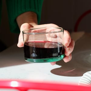 May include: A person holds a clear glass with a green base, filled with a dark red liquid, possibly wine. The person wears a green jumper. The image is lit by natural light, with a white surface in the background. The glass is a simple, elegant design.