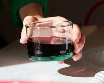 May include: A person holds a clear glass with a green base, filled with a dark red liquid, possibly wine. The person wears a green jumper. The image is lit by natural light, with a white surface in the background. The glass is a simple, elegant design.