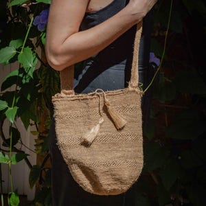 May include: A tan, woven shoulder bag made of natural fiber with a textured, open-knit appearance. The bag features a long, matching shoulder strap and a drawstring closure at the top, accented with two small, dangling tassels of the same material.