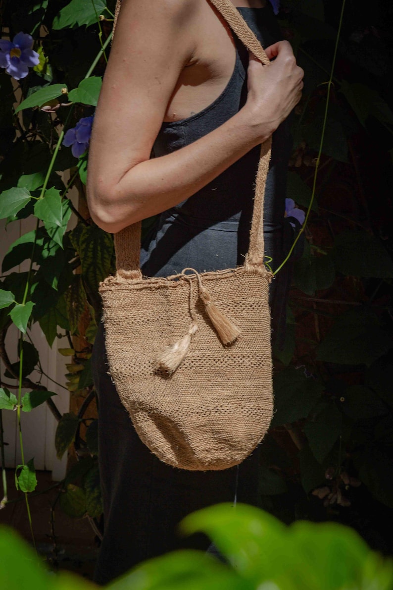 May include: A tan, woven shoulder bag made of natural fiber with a textured, open-knit appearance. The bag features a long, matching shoulder strap and a drawstring closure at the top, accented with two small, dangling tassels of the same material.
