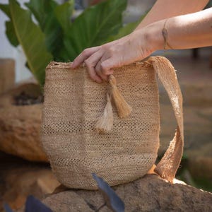 May include: A beige woven shoulder bag crafted from natural fiber, featuring a textured, open-weave pattern, a single wide strap, and two decorative tassels hanging from the top opening, showcasing a simple, unstructured bucket-style design.