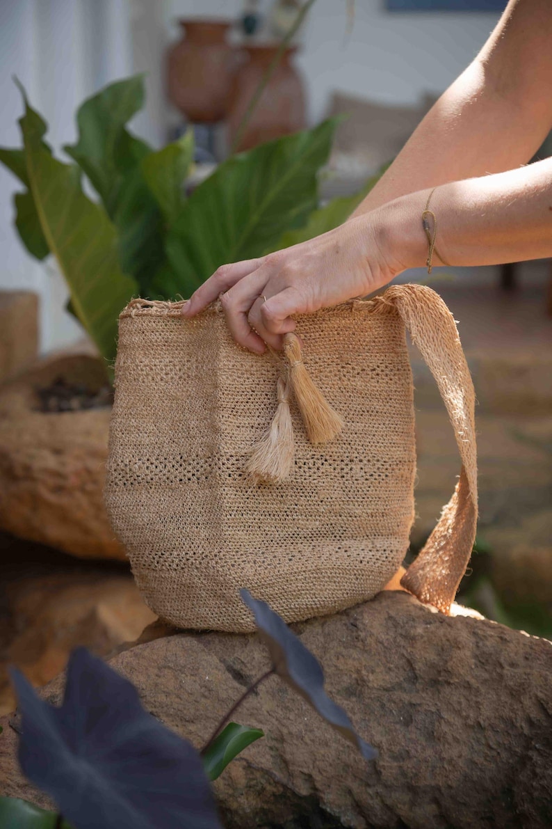 May include: A beige woven shoulder bag crafted from natural fiber, featuring a textured, open-weave pattern, a single wide strap, and two decorative tassels hanging from the top opening, showcasing a simple, unstructured bucket-style design.