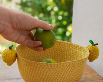 May include: A yellow woven basket with two decorative lemon-shaped handles. A hand is placing a green lime into the basket, which already contains another lime. The basket is sitting on a wooden surface.