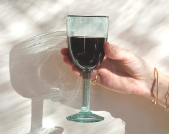 May include: A teal-tinted wine glass filled with red wine. The glass has a long stem and a wide base. The glass is held against a white background with shadows. The glass is likely made of glass.
