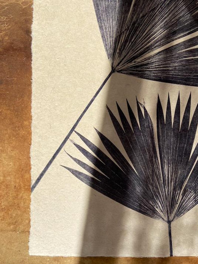 May include: A black and white print of two palm leaves on a cream background. The leaves are arranged in a symmetrical pattern, with one leaf on top of the other.