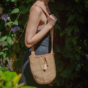 May include: A beige woven shoulder bag with a textured pattern, a single wide strap, and a small decorative tassel hanging from the top opening. The bag features a drawstring closure and a rounded bottom shape, crafted from a natural fiber material.