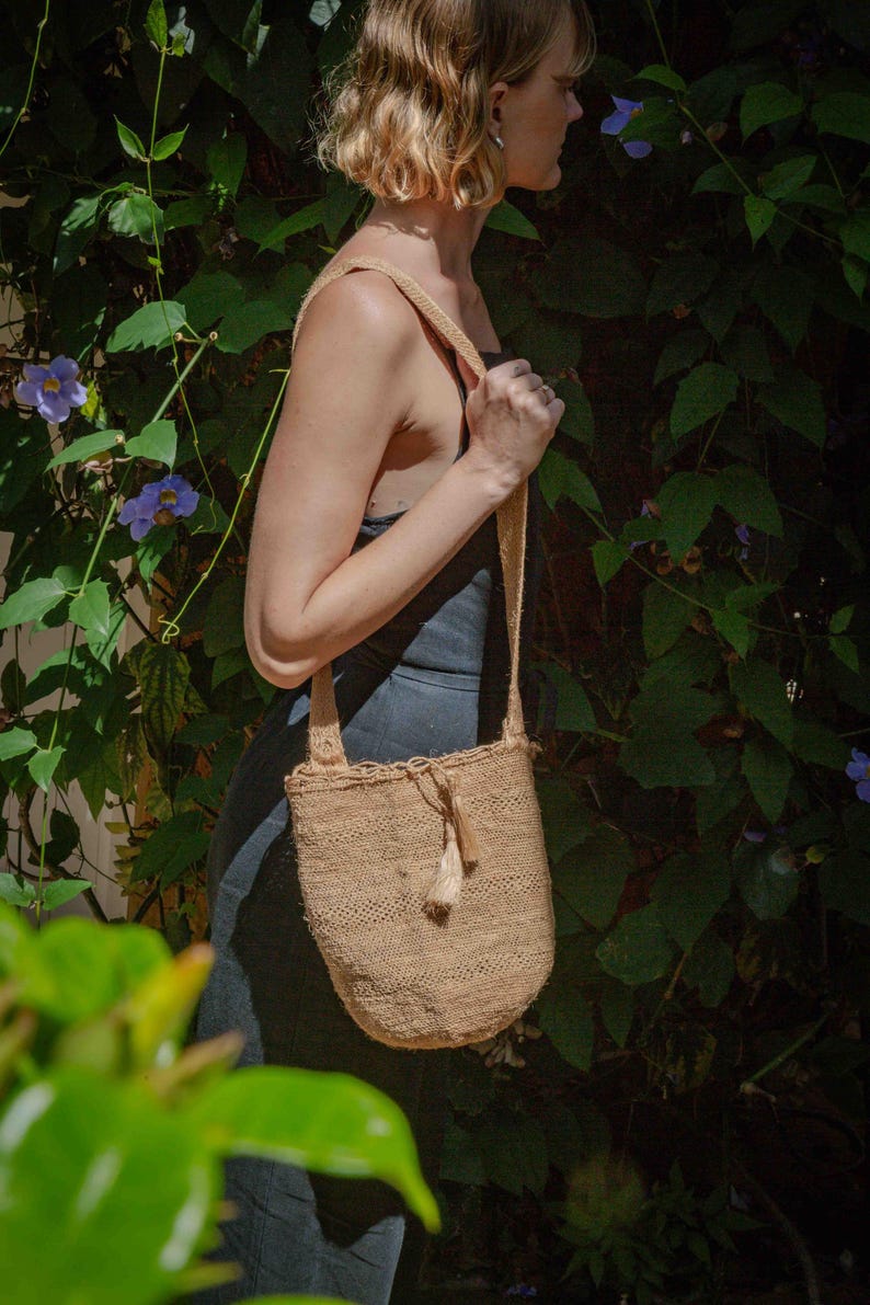 May include: A beige woven shoulder bag with a textured pattern, a single wide strap, and a small decorative tassel hanging from the top opening. The bag features a drawstring closure and a rounded bottom shape, crafted from a natural fiber material.