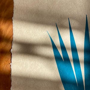 May include: A close-up of a teal blue print of a plant on a textured beige paper background. The print is of a stylized plant with long, narrow leaves.