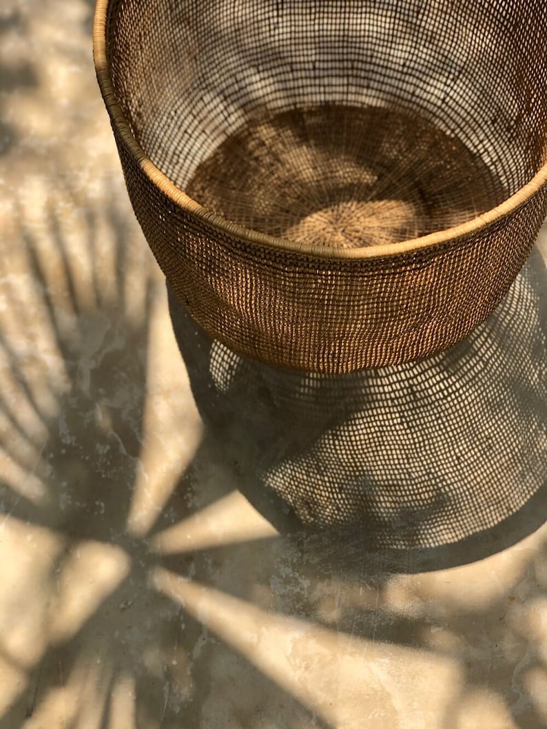 Op de afbeelding: Een grote, ronde, geweven mand gemaakt van natuurlijke materialen. De mand is leeg en heeft een lichtbruine kleur. De mand staat op een lichtbruine ondergrond.