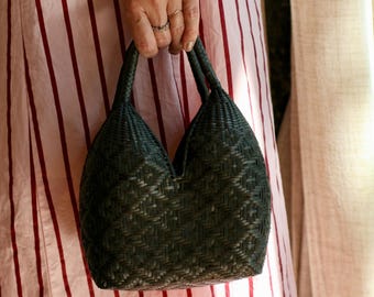 May include: A dark green woven handbag featuring a structured, rounded silhouette with two integrated handles and a V-shaped top opening. The bag displays a detailed, repeating geometric basket-weave pattern throughout its exterior surface.