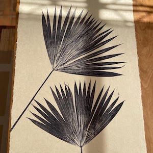 May include: Two black palm leaves are pressed onto a beige background. The leaves are arranged in a symmetrical pattern, with the tips of the leaves pointing towards the top of the image.