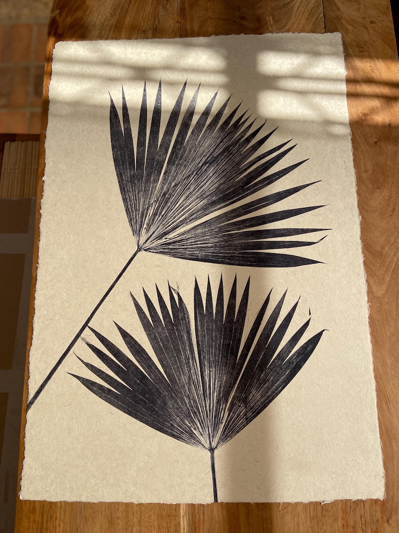May include: Two black palm leaves are pressed onto a beige background. The leaves are arranged in a symmetrical pattern, with the tips of the leaves pointing towards the top of the image.