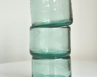 May include: Three clear, cylindrical drinking glasses stacked. The glasses have a slight teal tint and a rounded base. The glasses are made of glass and are on a white surface. Each glass is approximately 7 cm in height.
