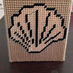 May include: A square box crafted from beige plastic canvas with a stitched design. The front panel features a shell motif created with light beige yarn and outlined in black yarn, showcasing a grid-like texture across the entire surface.
