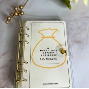 May include: A clear plastic binder with gold rings holds a white card with a yellow money bag illustration. The card reads "52 Money Pots Savings Challenge I am saving for..." and "Simple Money Plans".
