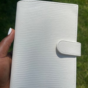 May include: White leather planner with a textured, embossed design. The planner has a white strap closure.