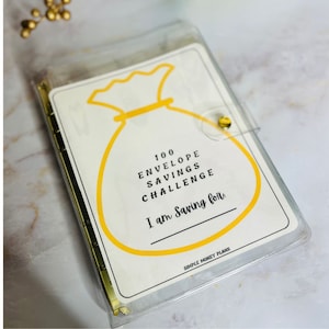 May include: A clear plastic envelope with a gold binder ring and a yellow money bag illustration. The text reads "100 Envelope Savings Challenge I am saving for:" and "Simple Money Plans".