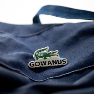 以下が含まれることがあります： 青いキャンバス地のバッグに、銀色のワニのロゴと白い文字で「GOWANUS」と書かれている。