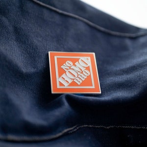 May include: A silver and orange enamel pin with the text "NO HOMO BRO" in white letters.
