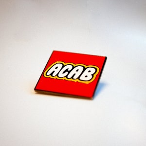 Peut inclure: Une brique rouge de style Lego avec l'acronyme "ACAB" en lettres blanches avec un contour jaune.