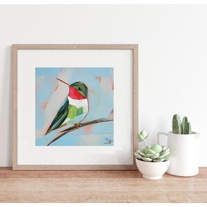 May include: A framed painting of a green, red, and white hummingbird perched on a branch. The background is a light blue color with white and peach brushstrokes. The painting is signed 'Dt' in the bottom right corner.