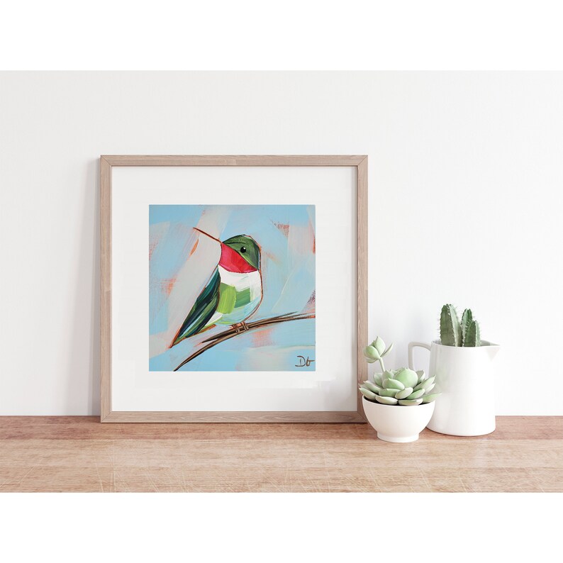 May include: A framed painting of a green, red, and white hummingbird perched on a branch. The background is a light blue color with white and peach brushstrokes. The painting is signed 'Dt' in the bottom right corner.