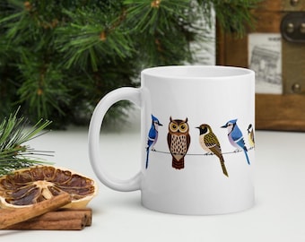 May include: White ceramic mug featuring a design of five colorful birds perched on a wire. The birds include a blue jay, an owl, a brown bird, another blue jay, and a small yellow bird. The mug has a white handle.