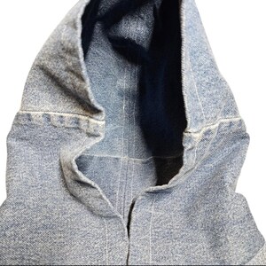 May include: Close-up of a light blue denim jacket hood. The hood is open, revealing a dark blue interior. White stitching details the seams and edges of the denim fabric. The jacket appears to be new.