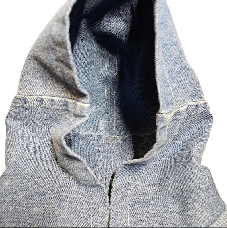 May include: Close-up of a light blue denim jacket hood. The hood is open, revealing a dark blue interior. White stitching details the seams and edges of the denim fabric. The jacket appears to be new.