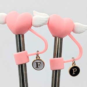 May include: Two pink heart-shaped straws with white wings and gold charms. The charms are round and have the letters "F" and "P" on them.