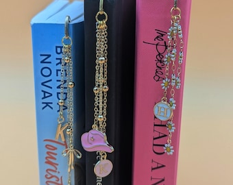 May include: Three bookends with different charms hanging from them. The charms are a pink cowboy hat, a letter K, and a white daisy with a letter H. The bookends are on a light brown surface.
