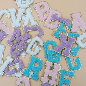 May include: Assortment of colorful felt letters with gold glitter trim. The letters are in various shades of pink, purple, blue, and white.