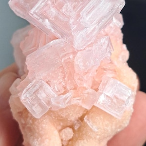 May include: A close-up of a mineral specimen featuring a cluster of translucent, cubic crystals in shades of pink and white. The crystals are stacked atop a light peach-colored base, creating a layered effect. The overall texture is rough and crystalline.