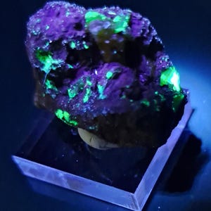 May include: A mineral specimen illuminated with a black light, showcasing vibrant purple and green fluorescence. The rock sits on a clear, rectangular display stand, creating a striking contrast against the dark background. The mineral's texture is rough and uneven.