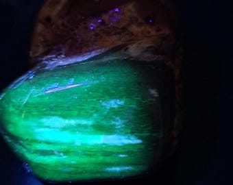 May include: A polished, rounded stone displaying a vibrant, glowing green luminescence across its smooth surface. The upper portion of the stone remains dark and textured, contrasting with the bright, neon-like green light emitted from the lower section.