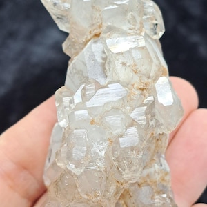 May include: A cluster of clear quartz crystals with multiple facets and a slightly translucent appearance. The crystals have a natural, rough texture and are held in a hand against a dark background. The quartz is a light, neutral color.