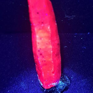 May include: A vertical, elongated mineral specimen exhibiting a vibrant, glowing neon red fluorescence under ultraviolet light. The crystal has a textured, uneven surface with small dark inclusions and is anchored to a dark, irregular base.