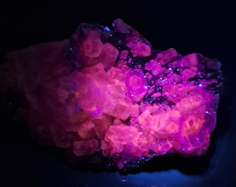 May include: A cluster of crystalline mineral formations glowing with intense neon pink and purple hues under ultraviolet light. The specimen features numerous angular, cubic-like structures that emit a vibrant, luminescent light against a dark background.