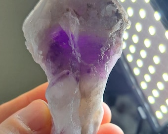 May include: A raw amethyst crystal with a mix of white and deep purple hues. The natural stone has a rough, uneven texture and is held in a hand. The crystal's color transitions from a pale white to a vibrant purple.