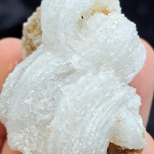 May include: Close-up of a white mineral specimen with a crystalline structure. The mineral has a layered appearance and sits on a beige matrix. The background is dark.