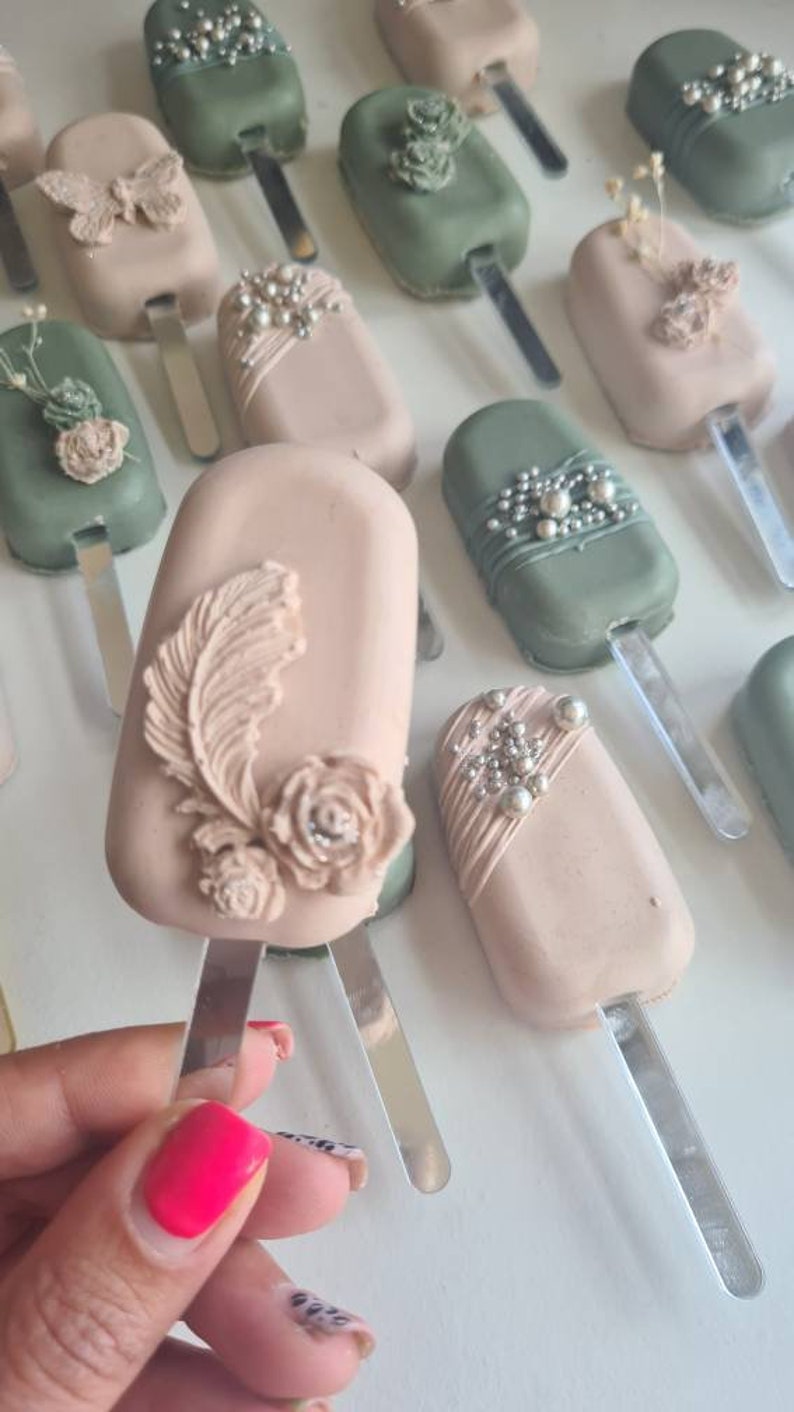 May include: An assortment of decorative, pastel-colored cake pops on sticks. Some are light pink and others are sage green, adorned with edible embellishments like pearls, feathers, and flowers. The pops are arranged on a white surface.
