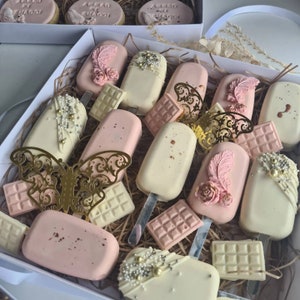 May include: A box filled with decorated cake pops and chocolate bars. The pops are pink and white, adorned with feathers, pearls, and gold accents. The chocolate bars are light pink and white. Some cookies are visible in the background.