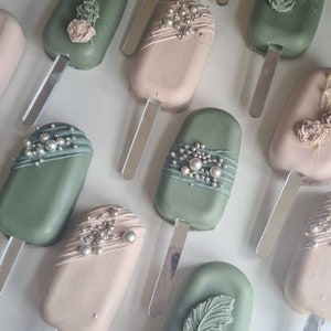 May include: An overhead shot of several pastel-colored cake pops on sticks. Some are sage green, others are light pink. The cake pops are decorated with edible pearls, flowers, and butterfly designs. The pops are arranged on a white surface.
